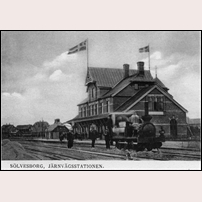 Sölvesborg, nya stationshuset byggt 1899. Bilden är tagen senast 1902. På taket vajar unionsflaggan. Loket är ett av banans engelska maskiner från 1870-talet med den speciella utformningen av förarhytten. Foto: Okänd. 