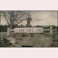 Gräns mellan Skåne och Småland på ett privat vykort postgånget 1930. Bron är den gamla riksvägens korsning med gränsbäcken och skylten är avsedd att läsas från järnvägen som löper parallellt med landsvägen på 50 meters avstånd. Foto: Okänd. 