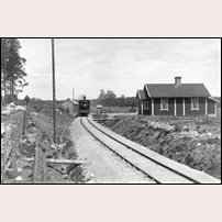 5 Rockneby 1898. Banan är nyöppnad och ett av de första loken, nr 3 Frigga, drar ett tåg söderut förbi stugan. Den kvinnliga grindvakten har stängt grindarna över landsvägen och tittar ut ur den lilla kuren medan tåget passerar. Bild från Järnvägsmuseet. Foto: Okänd. 