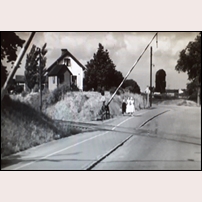 711 Bulltofta sommaren 1943. Banvaktshustrun har från vaktkuren påbörjat fällningen av vägbommarna och står utanför och övervakar att allt fungerar som avsett. Bild från samma SJ-film som den föregående bilden. Foto: Okänd. 