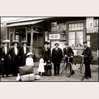 Holsby kombinerade stations- och banvaktsstuga troligen på 1910-talet. De ståtliga hattarna och de långa kjolarna damerna bär visar att bilden är tagen innan 1920-talets mer uppsluppna kläd- och hattmode kom i bruk. Foto: Okänd. 