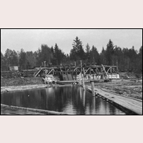 Östbyberg lastplats med timmeruppfordringsverk 1906. Lastning pågår, några vagnar står färdiglastade till vänster, några tomvagnar ses till höger och ett antal stockar är halvvägs upp på elevatorn. Inemot tio personer är sysselsatta med lastningen. Foto: Okänd. 