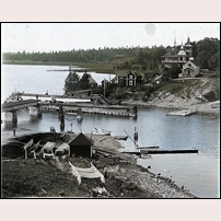 Järnvägsbron över Mälaren vid Kvicksund före 1924. Bilden som är tagen i nordvästlig riktning visar järnvägens första svängbro, här i öppet läge. På norra sidan av vattnet ses bryggan för vägfärjan. Motsvarande brygga på den södra sidan syns dåligt men den branta vägen ned till bryggan visar platsen. Snett till höger ovanför svängbron ligger banvaktsstugan Kvicksund, här i påbyggt skick. Kvicksunds hotell är huset med tinnar och torn längst till höger.  Foto: Okänd. 