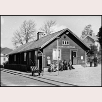 Ljusfallshamnmar station den 25 maj 1961. Stationshuset finns kvar, ännu i full tjänst. Skylten för Postverket och Postsparbanken pryder fasaden, och dörren till väntsalen står öppen. Människor samlas vid stationen, för att vänta på tåget eller bara få en pratstund. Bagagekärran är redo att transportera smågods fram till rälsbussen. Järnvägen är fortfarande bygdens ryggrad, och posten är dess nervsystem. Men i bakgrunden finns den som ska komma att förändra allt, bilen. Bild från Järnvägsmuseet. Foto: Okänd. 