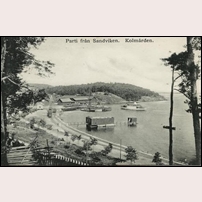 Kolmården station 1910, "sedd från väster med Serveeringsberget i fonden. Tågsätt framför stationshuset, loket gör rundgång på det andra spåret. Båten på ingående. Man ser alla gamla skjul vid bryggan liksom de "avskurna" magasinen  mot linjen som försvinner till vänster i bakgrunden. Järnvägen har planterat bangårdsområdet i förgrunden med bl.a. träd" (så långt är texten hämtad ur Lennart Welanders fina bok om Nunnebanan). Till höger går spåren ett par hundra meter längs stranden och slutar med en vändskiva framför det lilla lokstallet. Okänt vykort. Foto: Okänd. 